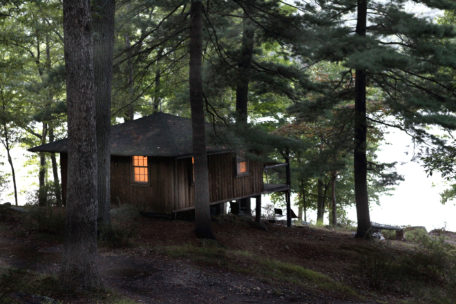 Baker Camp shuts down in the late fall after a summer of outdoor tourism. A cabin is perched in the trees at nightfall, with a single light on in the window, on the shores of Sebago Lake in Harriman State Park.